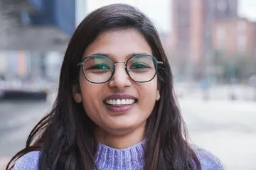 Close-up portrait of a smiling young South Asian woman with glasses and a purple sweater, showing a healthy white smile in an outdoor city setting for a customer review.
